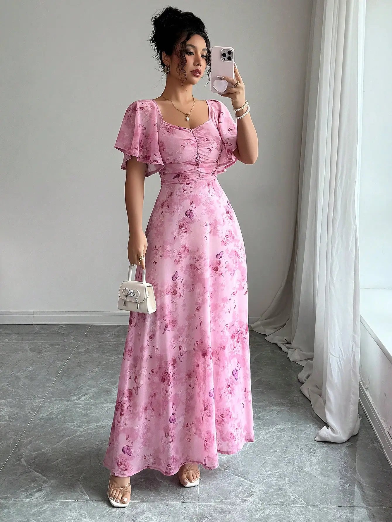 Woman taking a mirror selfie wearing a pink floral dress in a room with white curtains.